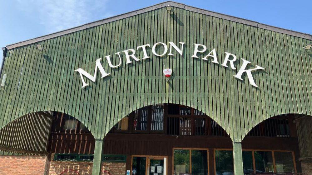 An exterior shot of a large red brick, barn-style building, with a green wooden awning and white lettering sign that reads Murton Park.