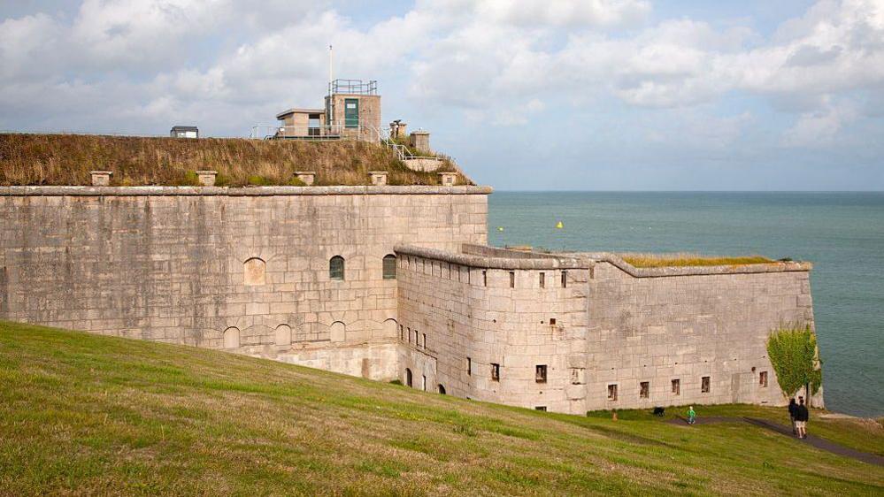 A photograph of the stone walls of Nothe Fort taken from afar, showing a section of the perimeter. A green hill rolls down to the sea in the foreground, with the sea in the background.