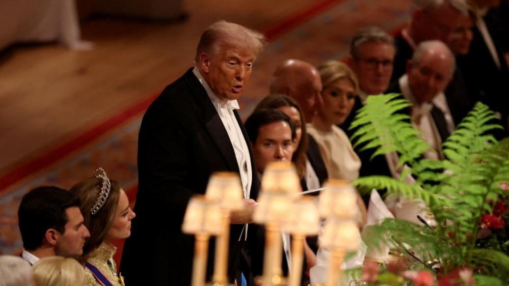 Donald Trump in a black dinner suit stood up addressing the dinner guests. In the foreground, but out of focus, is a lamp shade and a green fern.