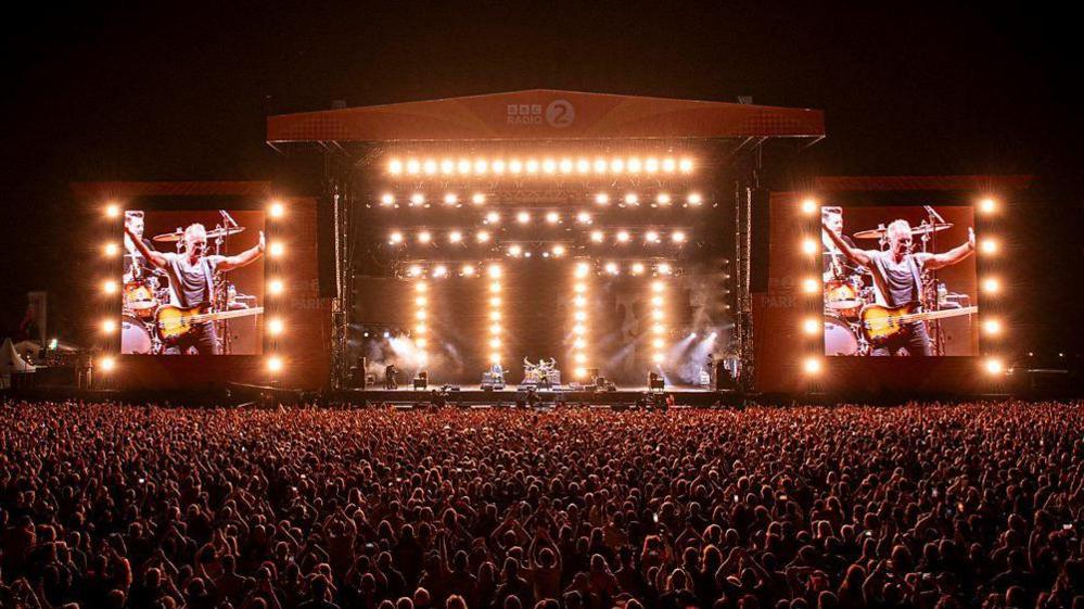 The BBC Radio 2 in the Park stage, as well as a large crowd standing in front of it, is illuminated by bright lights. 