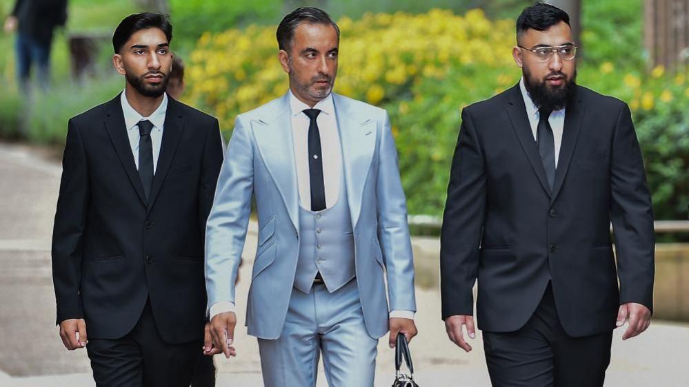 Three men walk towards the camera. The two men on either side are wearing black suits and ties and white shirts and the man in the middle is wearing a light blue three-piece suit and holding a briefcase.