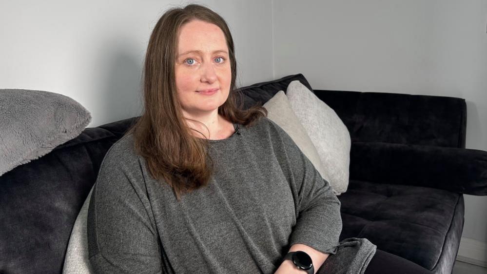 Amy Simpson sitting on a sofa, looking at the camera, with long dark hair, wearing a grey top, with a black watch on her wrist. The sofa is dark and the walls are grey.