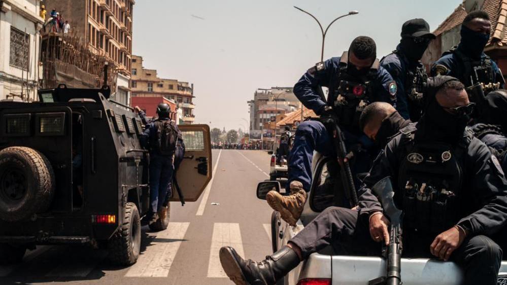Security forces sit in the back of a truck, and there is a bear cat vehicle next to them. Many protesters can be seen in the distance.