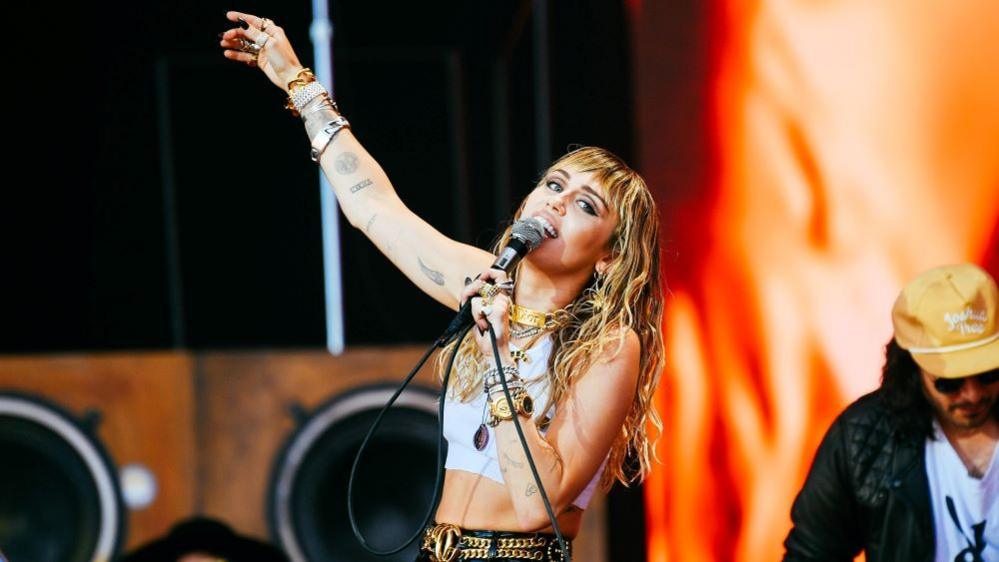 Miley Cyrus wearing a white cropped tack top and gold bracelets performing on the Pyramid Stage at Glastonbury Festival 2019 