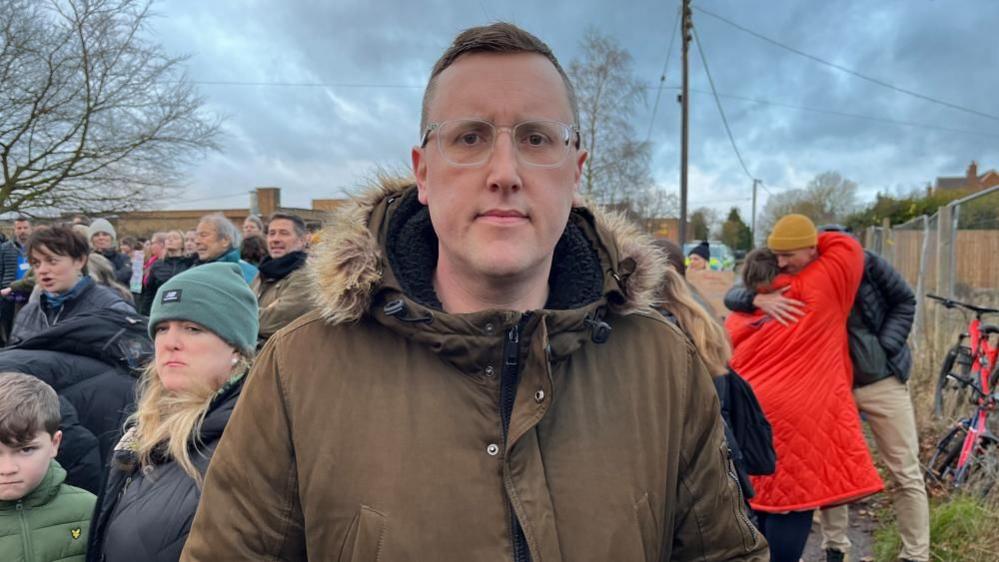 Scott Lyons, Norfolk branch secretary of the National Education Union, wearing a brown coat with fur lined hood. He is standing outside Reepham High School and College with parents and pupils on a protest about redundancies.