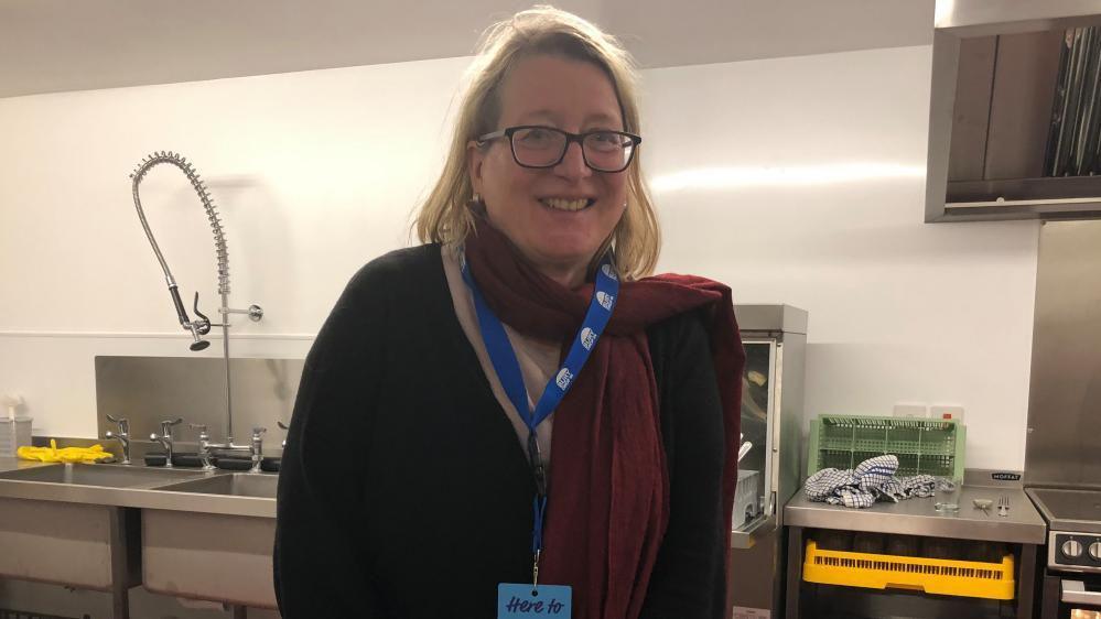 Sabine Dornbusch is standing in a kitchen hatch. She is wearing a dark jumper and has mid length blonde hair. She is standing in the kitchen and in front of a serving hatch. 