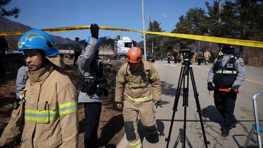 Firefighters and security personnel crossing under a yellow police tape cordoning off the bomb site