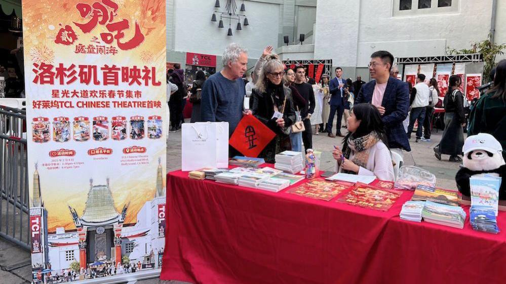 A standing banner announcing the premiere of Ne Zha 2 in Los Angeles. Beside it is a table covered by a red cloth. An Asian woman sits behind the table, which has stacks of promotional brochures. The woman is speaking to a white couple. 