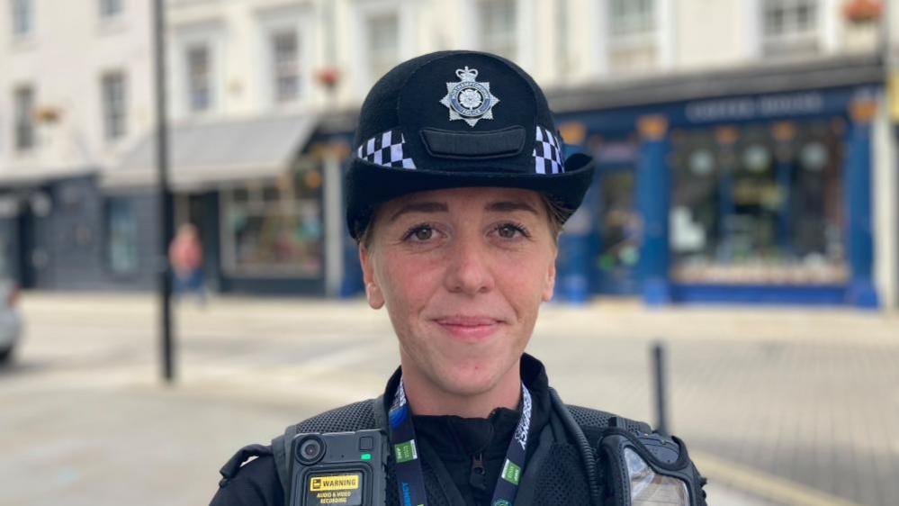 A female police office stood on a street with shops behind her. She is looking at the camera and smiling.