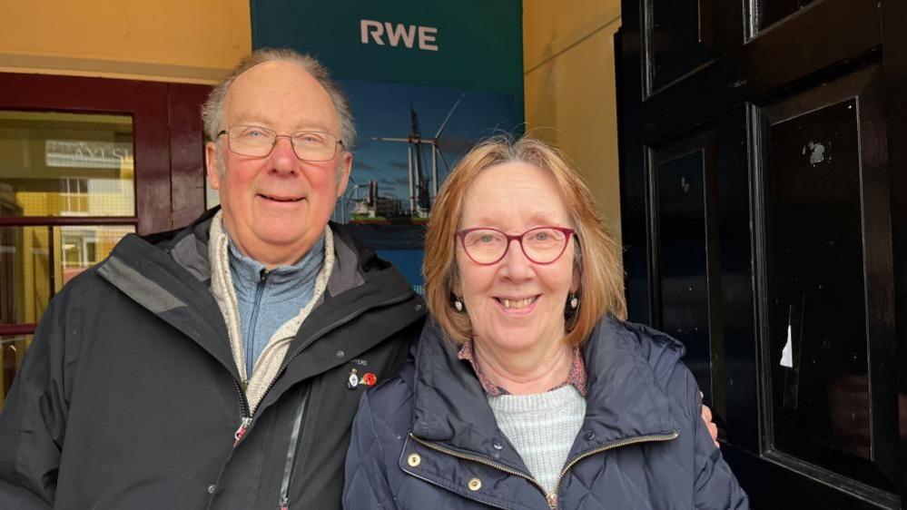 Dave Eccles stands on the left of his wife. He is wearing a black coat, with a white fleece and blue zipped shirt beneath that. He has glasses and receding grey hair. His wife, Vivien, has blonde hair and a centre parting, is wearing red-rimmed glasses and has earrings. She is wearing  a blue jacket and grey jersey beneath. They are standing near an RWE banner inside the entrance to Aylsham Town Hall.