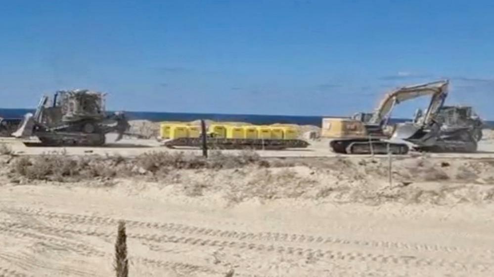 Two excavators are pictured on either side of a set of yellow blocks on sandy ground in Gaza