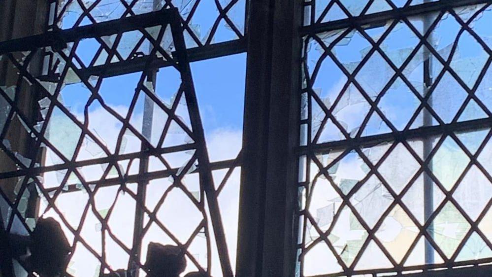 Several diamond panes are smashed with leadwork warped and damaged. The sky can be seen outside.