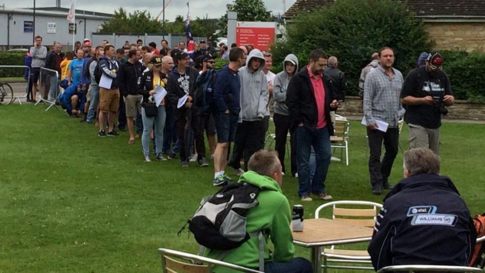 A queue of people, some in grey hoodies and some in shorts, is making its way towards the camera. There are grey buildings in the background. The queue of people is standing on grass. There is a small table in the foreground at which two people, one wearing black and the other wearing green, are sitting.