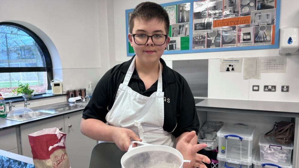 Logan, a young boy, with glasses on, with short dark hair, wearing a black T-shirt, with a white apron. There is a sink, kitchen equipment behind him, and pictures on the wall. Boxes are to the right. He is holding a sieve. 