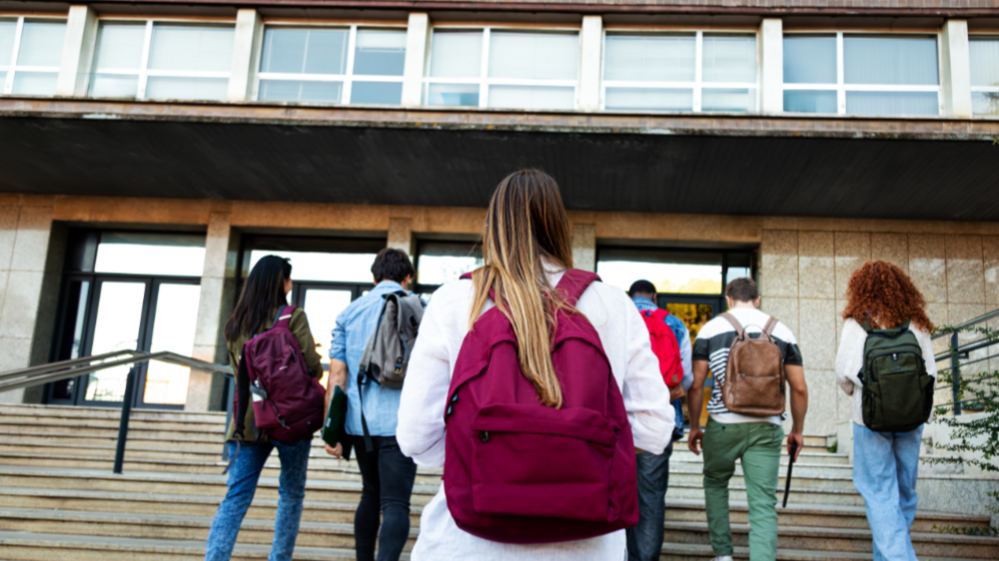 A stock image of university students walking up some stairs into a university building. A young woman is at the forefront of the image, she has long blonde hair and is wearing a maroon backpack