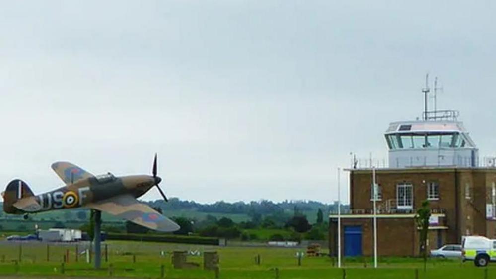 A statue of a spitfire is next to a brick control tower at the airfield. Behind them are rolling fields.