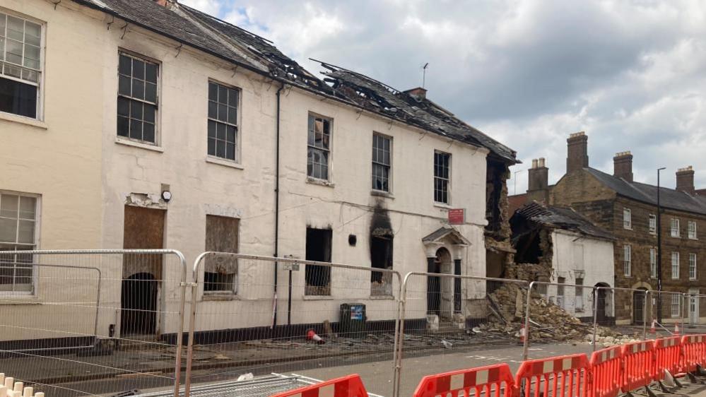 Two white two-storey houses next to each other. Part of the roof is missing. Some of the windows are broken or boarded up. Both houses have medium-sized windows made up of 12 squares. There are scorch marks above one of the windows. The small red Ex-servicemen's Club sign is still visible over the Georgian doorway.