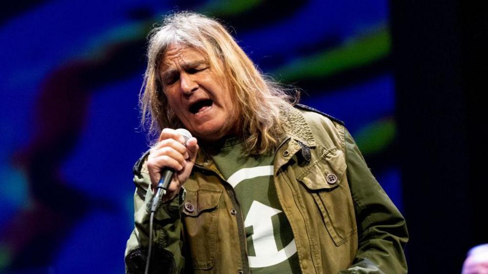 Mike Peters on stage singing. He has long hair and is wearing a green jacket and a green t-shirt.