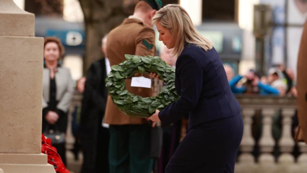 O'Neill holds a green wreath as she moves to place it at the base of a Cenotaph where a poppy wreath already lies