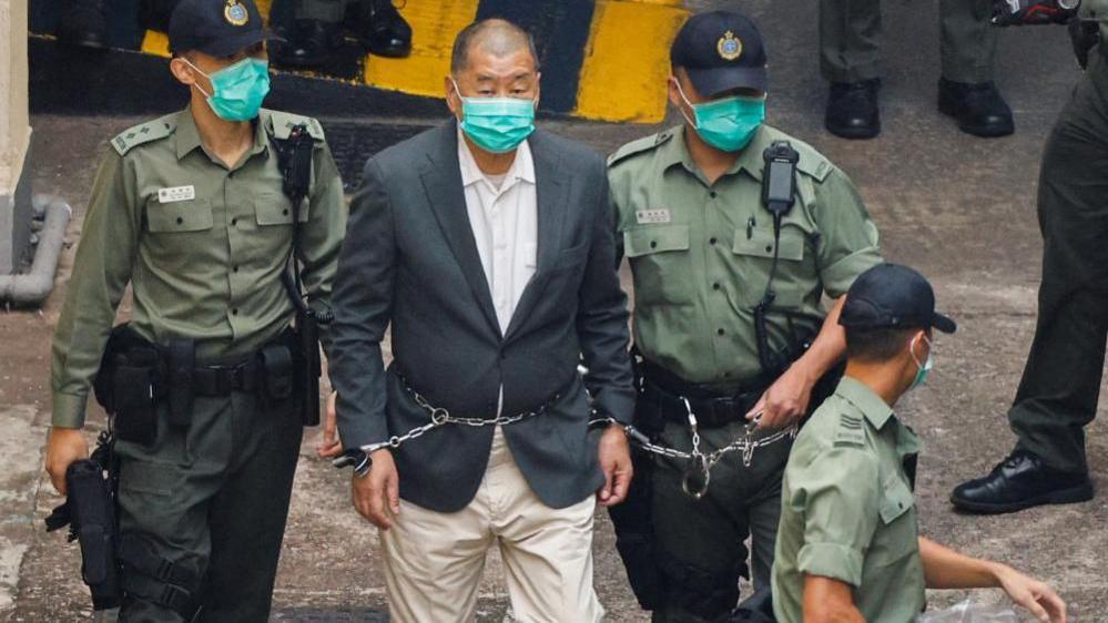 Lai, dressed in a gray suit and beige pants, walks handcuffed and flanked by three police officers to a police van