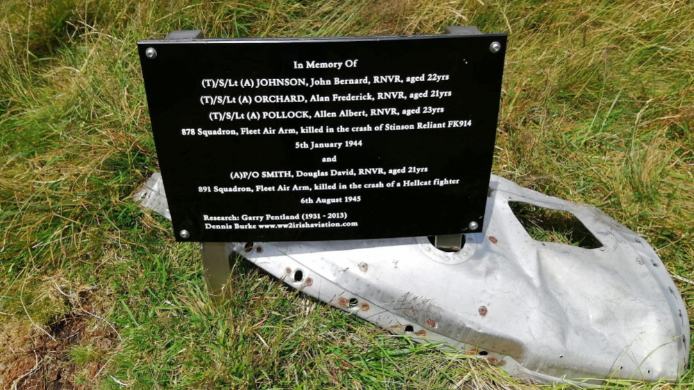 A memorial for the Petty Officer, on a black plaque with white writing, it sits above a small silver piece of wreckage. The sign states the names of those killed in the crash and their ages.