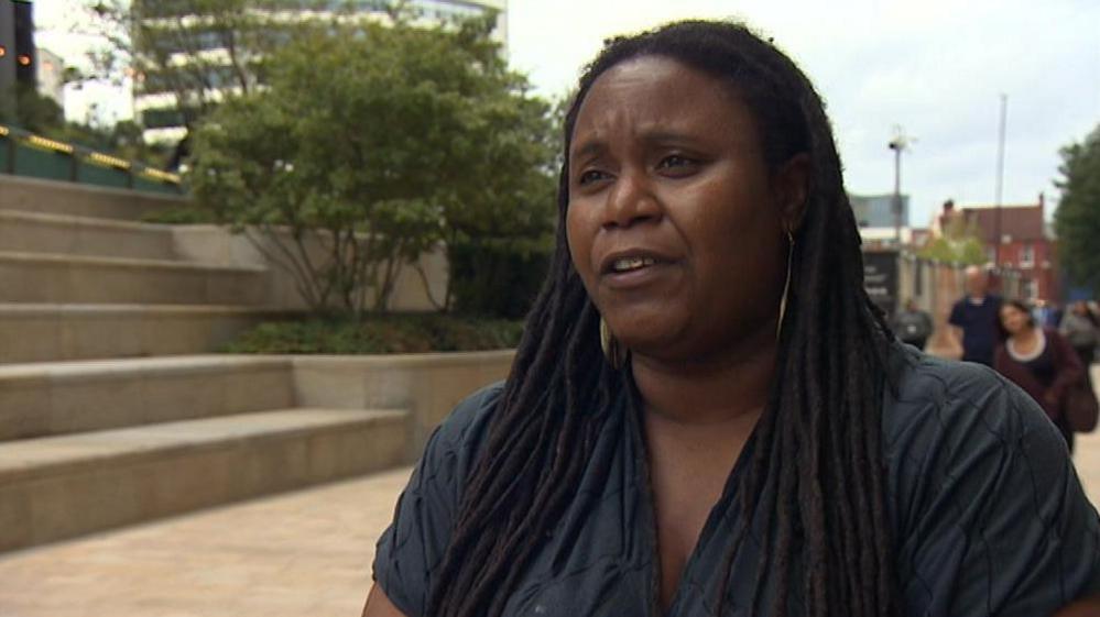 Sharon Thompson giving an interview. She has dreadlocks and is wearing a blue dress and earrings. We can see the area around the Paradise Estate and The Octagon tower behind her. There are people walking up and down but they are blurred out. 