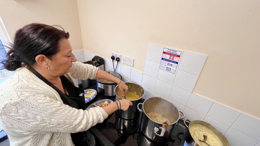 Joanne Beek is standing at a kitchen counter stirring food in a large metal pot. Several other pots and a slow cooker are on the black counter too. 
Joanne is wearing a cream-coloured knitted cardigan with a black apron  on over a black top. 