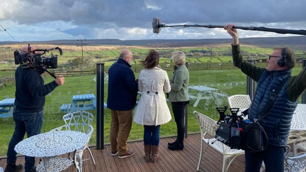 Two men and a woman stand on a restaurant patio overlooking the moors of Yorkshire. Two men with cameras and a boom microphone are filming them. 