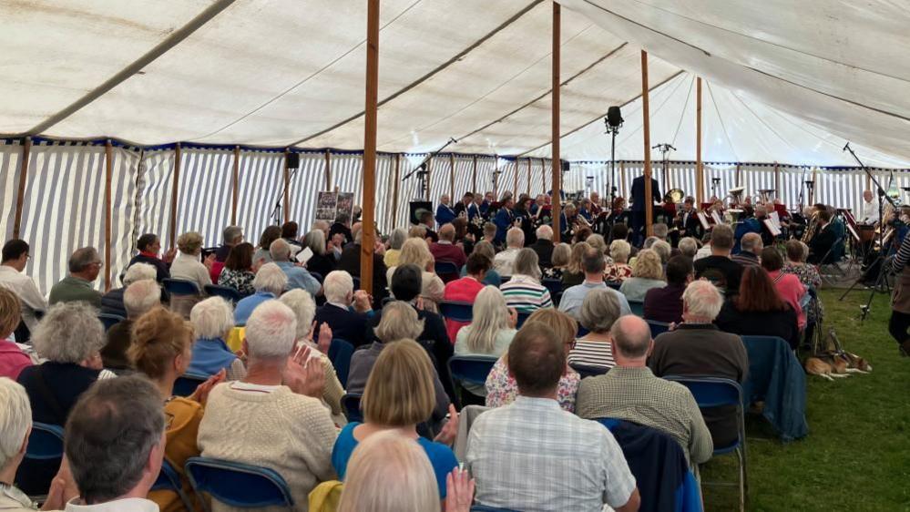A marquee with about 200 people sitting on folding chairs, looking at a brass band performing in front of them.