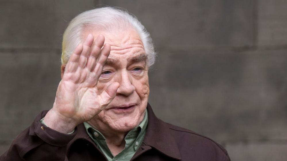 Brian Cox looking over to the right waving to someone. He is smiling and wearing a green shirt and brown jacket.