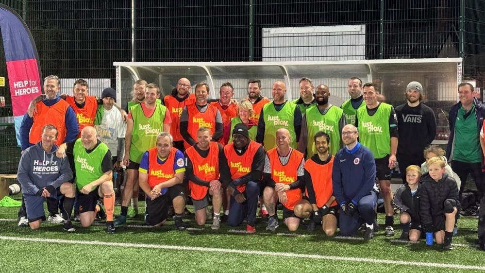 A group of football players wearing high visibility bibs in orange and green stand together as a team by the dugout on the side of the pitch. Former England player Emile Heskey is in the centre.