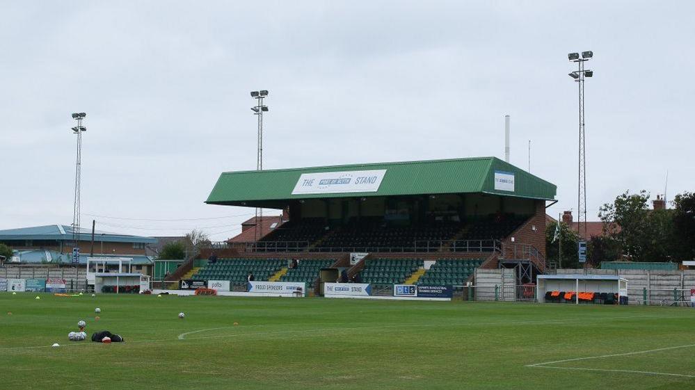 Croft park which has a small, green covered stand in the distance. There are floodlights on either side and buildings behind the stand. The pitch sits in front with various balls and cones in the centre.