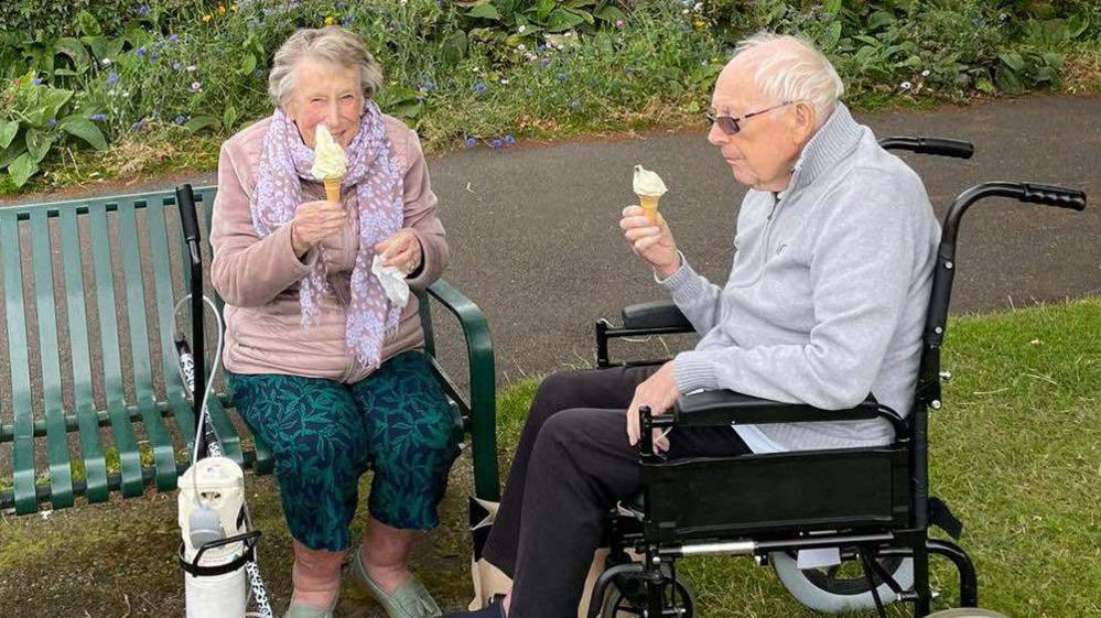 An elderly couple sharing ice creams sat on a bench outside, with the man sat in a wheelchair