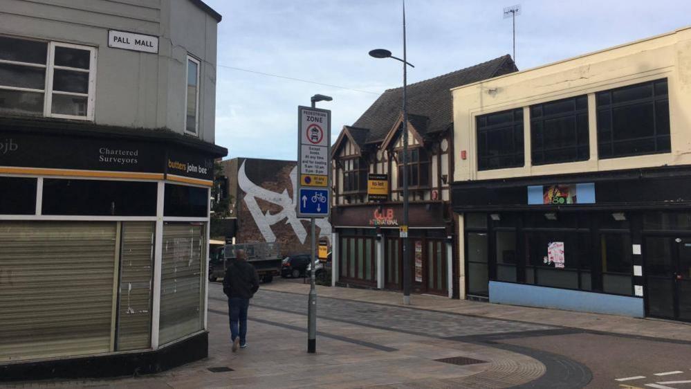 A shopping street that has several empty buildings. A man is walking away from the shot and there is a "to let" sign on one of the empty buildings on the right. The sky is a cloud but still pale blue. The road sign reads "Pall Mall". 