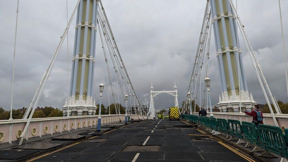 A view from the middle of the road across Albert Bridge. The clouds are thick and quite dark. There are buildings and trees and and a highway maintenance van in the background.