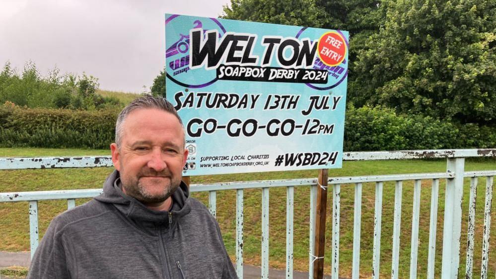 Man in a grey hoodie standing by a blue sign advertising the Welton Soapbox Derby