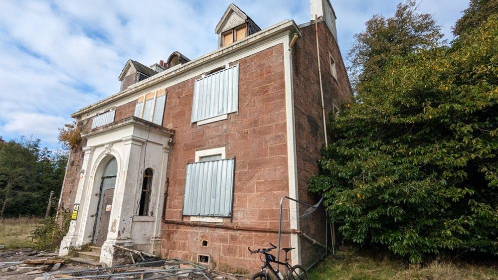 Georgian country house Carsebridge House, a two-storey red sandstone manor with a dark roof and white details around the symmetrical windows and doors. It is in a state of disrepair with boarded up windows and debris littered outside the front of the building. Trees and grass have grown around the building.