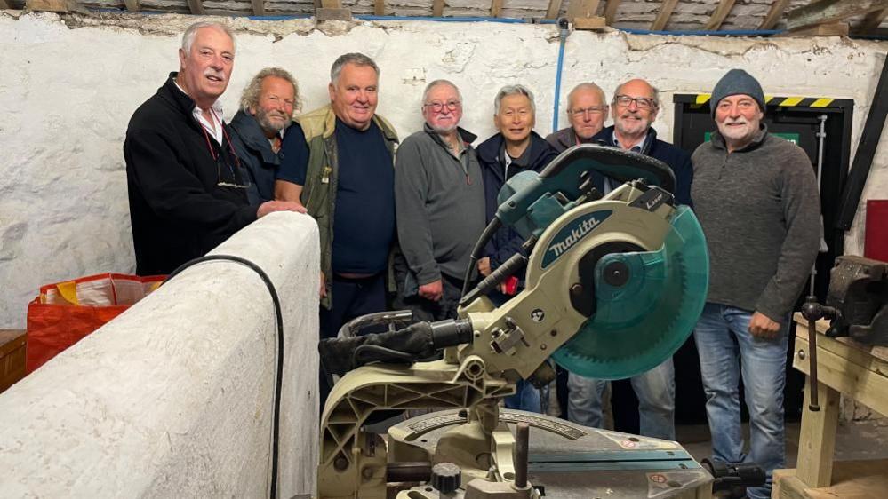 A group of men standing in a shed with white walls, behind a large circular saw. One of the men is wearing a green woolly hat.