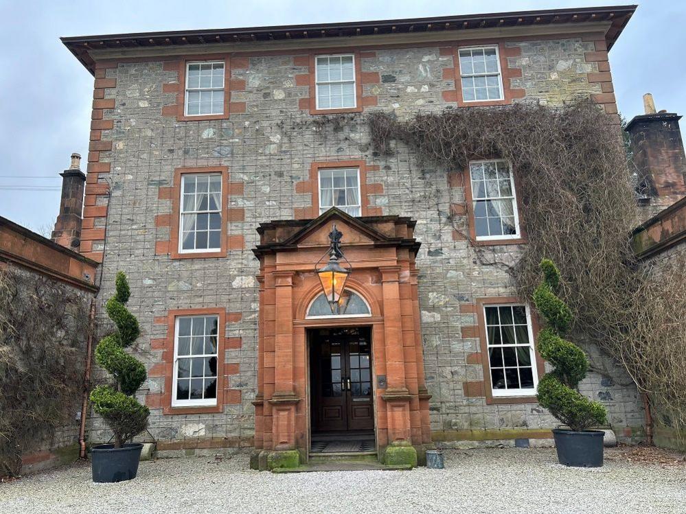 A large three storey sandstone building. There is gravel to the front and topiary on either side of a grand front door. 