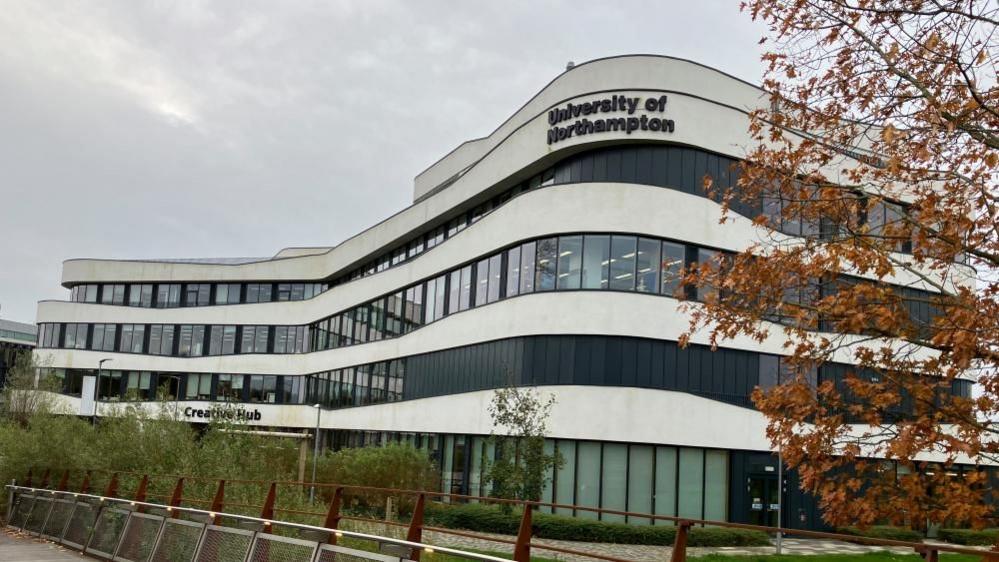 The exterior of the University of Northampton's waterside campus - a curved white and navy building with lots of glass windows overlooking a bridge.