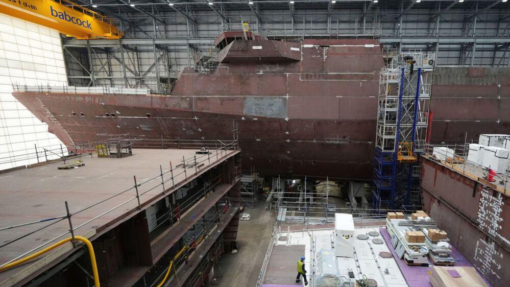 Construction work on Royal Navy's Type 31 warships in the Venturer building at Babcock International Group, in Rosyth. One warship is under construction with scaffolding around it. It is a rusty brown colour. The ship towers above a workman in a hi vis jacket and hard helmet below.