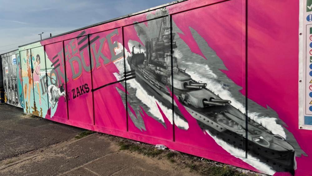 Hoarding painted pink with a silver-grey ship, depicting HMS Iron Duke. Some text is painted: THE IRON DUK, BY ZAKS. Images bleeding from elsewhere in the hoarding are visible on the extremes of this picture.