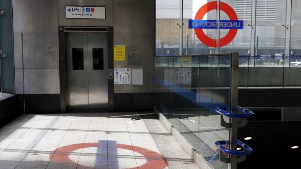 The ground-level indoor entrance area of a Tube station. On the left, there is a lift entrance with a sign above reading 'Lift to Underground', and on the right there is a set of stairs going down. Behind the stairs, there is a glass window with a Transport for London roundel on it. The roundel is also visible reflecting onto the ground in front of the lift.