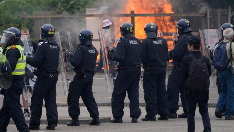 A line of riot police holding large shields, while a man walks past a generator, with fire burning in the background.