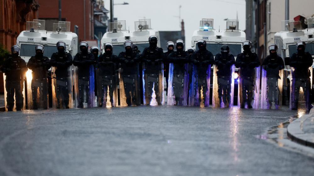 A line of riot police in front of police land rovers. They are wearing full protective gear, including helmets and protective shields.