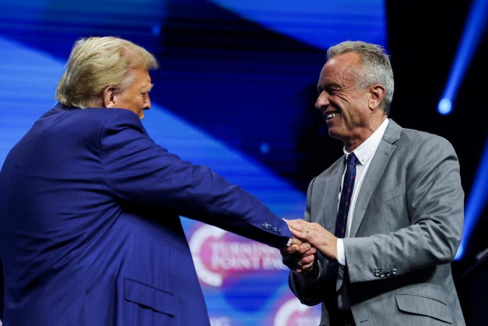 Donald Trump seen shaking hands with Robert F. Kennedy Jr. Trump is wearing a blue suit and has his back slightly to the camera. Kennedy is wearing a grey suit and smiling.