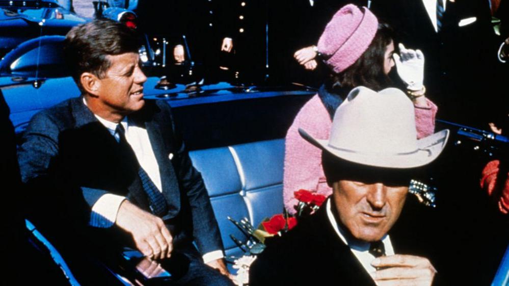 Kennedy seen in the backseat of an open convertible seated next to his wife who is wearing a pink outfit. A man in front of him is wearing a white cowboy hat