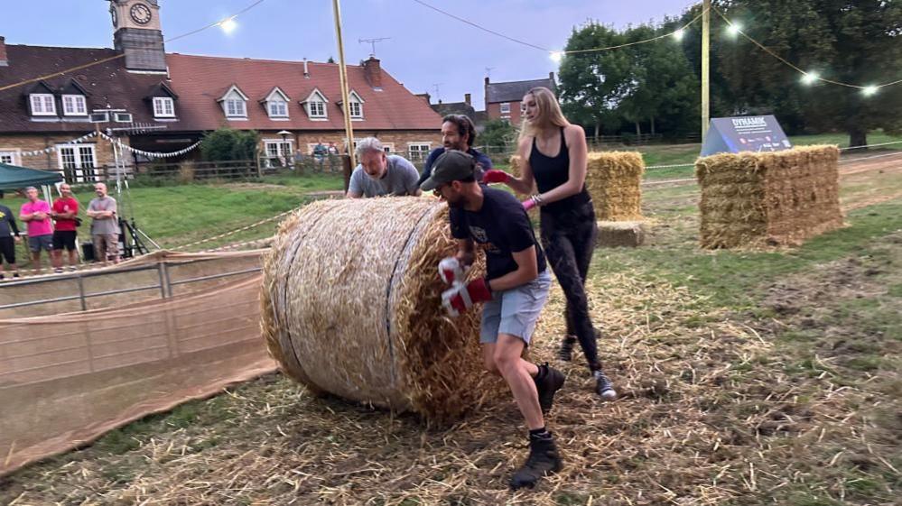 Three men and one woman push a large straw bale around the 180 degree bend at the end of a grass course. A few spectators are visible to the left. A row of cottages can be seen in the background.  It is starting to go dark and a string of lights is illuminating the scene.