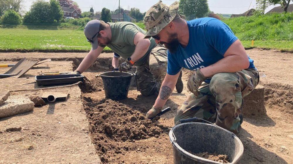 Two men in a shallow dry trench digging with trowels with archaeological equipment