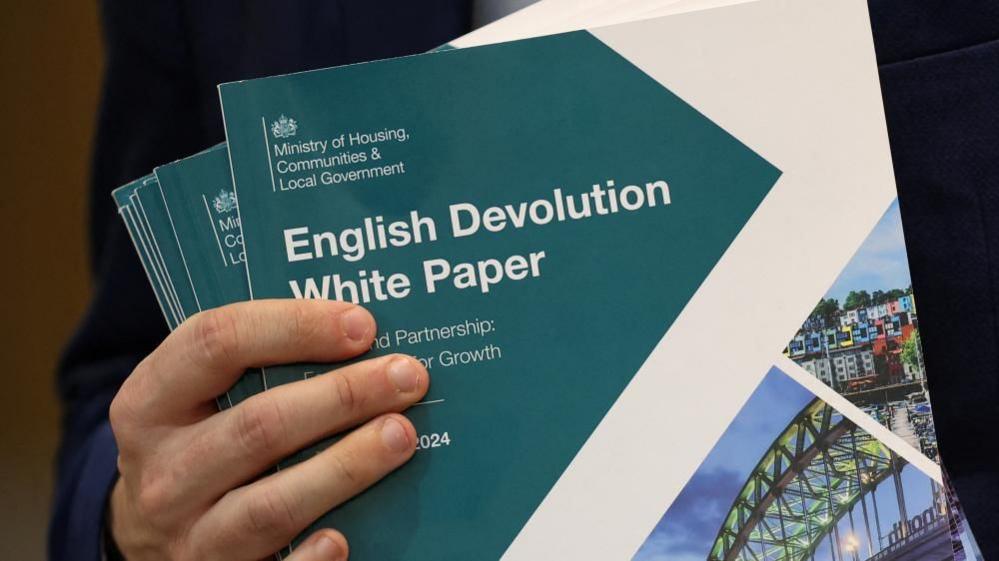 The 'English Devolution White Paper' is visible. It is a green and white booklet. Someone is holding several copies. Only their fingers are visible, wrapped around the booklets.